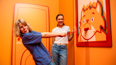 Zwei Frauen vor orangefarbener KatzeQ-Wandgrafik, ziehen an einer Tür im Escape-Room-Stil.